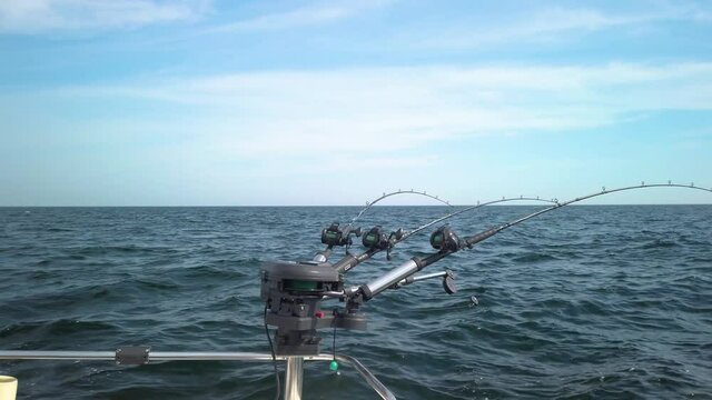 Three fishing rods hooked up with spinners, rigged for fishing, with lines out in the water. The rods are lined up in a row on a small fishing boat. Gentle waves makes the boat rock. 