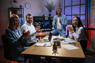 Group of four multicultural company workers sitting together at office desk, smiling on camera and clapping in hands. Male and female colleagues having working meeting and using gadgets.