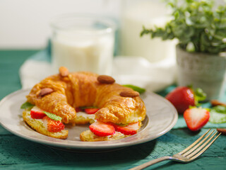 Close-up. Nicely served breakfast table for one person. Fresh French croissant with strawberries, almonds, mint, fork. In the background there is milk and an indoor flower.