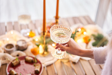 Hand with a glass of champagne in the foreground. A small table is served with snacks, bruschettas, and canapes. A decorated dining table with champagne glasses, candles and christmas tree an garland