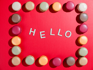Multicolored macaroon cookies. Pink, green, coffee, and red on red background with Hello wooden inscription 