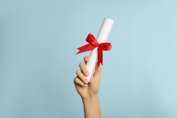 Student holding rolled diploma with red ribbon on light blue background, closeup