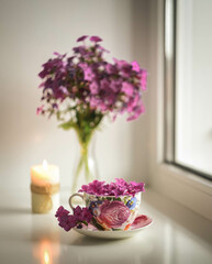 flowers are in a tea mug, a bouquet of flowers in a vase and a burning candle are in the background, a light background