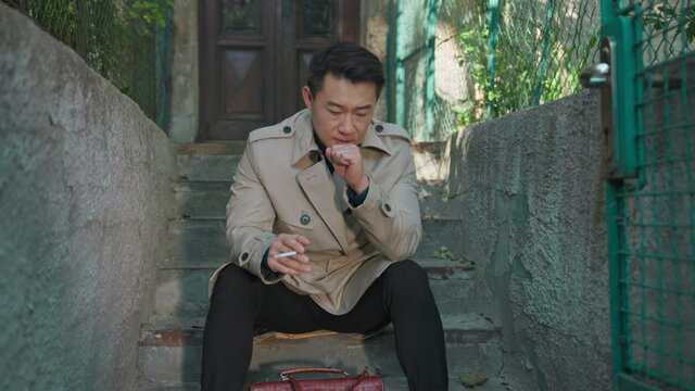 Asian frustrated businessman in suit coat smoking cigarette on stairs terrace outdoors. Handsome rich man in depression. House background.