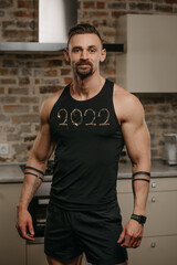 A kind muscular man with a beard is posing in his apartment after a workout. An athletic guy with tattoos on his arms is demonstrating his shirt with 2022 at home. A smartwatch is on the wrist.