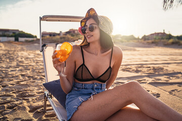 Beautiful young woman drinking an orange aperitif standing sitting on a chaise longue on the beach...