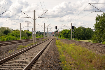 Fototapeta na wymiar Railroad tracks in the green nature. Steel tracks, light signs and electric poles. Beautiful white clouds on the overcast sky on a summer day. Empty railways heading to a small village.