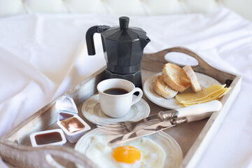 A breakfast tray with a fried egg, a cup of black coffee and a coffee pot are on a white bed. Retro style. Free space for text.