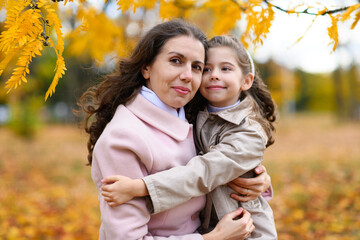 Fototapeta premium Mother and daughter portrait in an autumn park. Happy people pose against the background of beautiful yellow trees. They hug and are happy together.