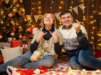 Portrait of a couple in New Year decoration. they show a heavy metal gesture - goat horns. Festive lights, gifts and a Christmas tree decorated with toys.