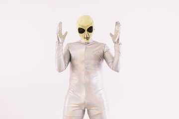 Person dressed in silver suit and green alien mask raising hands in surprise, over white background