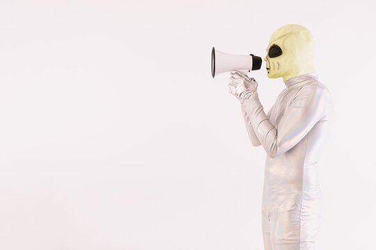 Person dressed in silver suit and green alien mask, speaking with a white megaphone, on white background