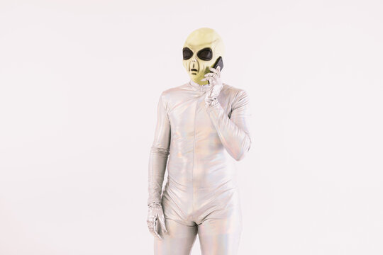 Person dressed in silver suit and green alien mask talking on the mobile phone over white background