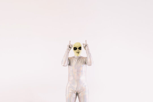 Person dressed in silver suit and green alien mask, pointing with his hands and fingers up, on white background