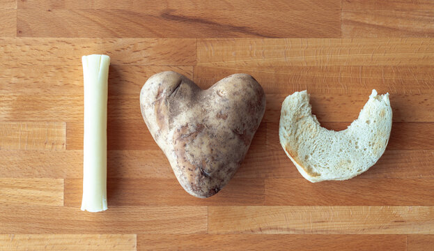 I Love You Message Spelled Out Using Food Items Including A Mozzarella String Cheese Stick, Heart Shaped Potato And An English Muffin Half With A Bite Out Of It On A Cutting Board Background.