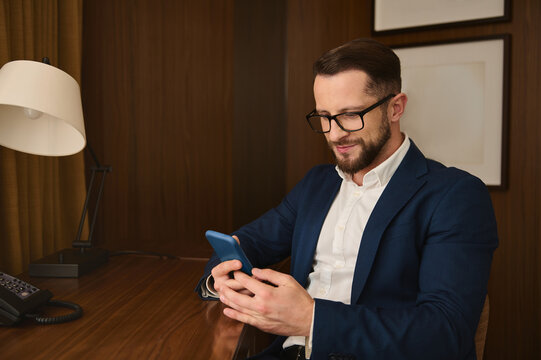 Handsome prosperous successful businessman, business traveler, investor wearing eyeglasses, smiles, using a mobile phone sitting at a table while resting in hotel room during business trip