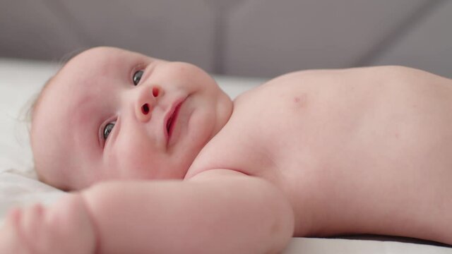 Happy Beautiful newborn baby, closeup face. Cute tiny baby with bare belly looks up, slow motion. Active newborn boy lies on the bed and moves his arms. Low angle view of a chubby baby..