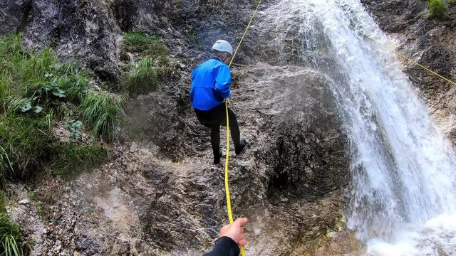 Point of view shot of a men helping a girl in a blue and black wetsuit rappel down a waterfall.