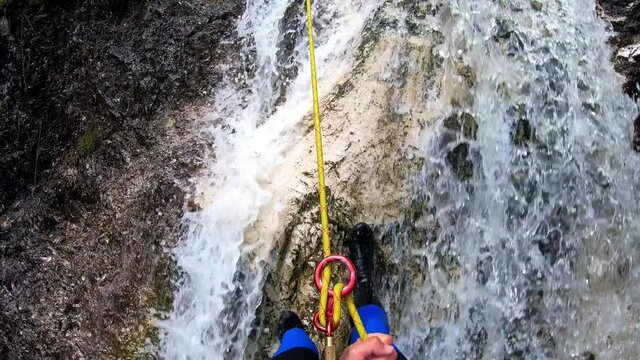 Point of view shot of a men rappelling down a waterfall.  He is wearing a black and blue wetsuit.