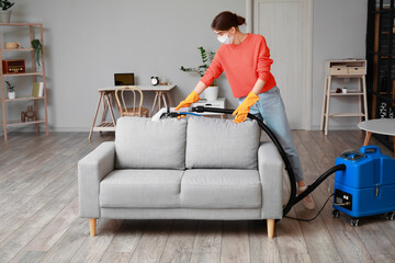 Fototapeta na wymiar Woman removing dirt from grey sofa with vacuum cleaner in room