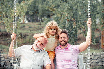 Boy son with father and grandfather swinging together in park outdoors. Three different generations ages grandfather father and child. Dad, son and granddad hugging.
