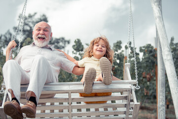 Child boy and grandfather swinging in summer garden. Grand dad and grandson sitting on swing in...