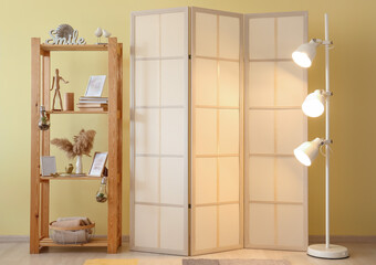 Folding screen with glowing lamp and shelving unit near yellow wall