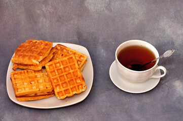 Several fresh Viennese waffles on a plate and a cup of black tea on a gray background.