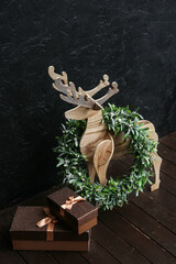 Wooden reindeer with Christmas wreath and gift boxes near dark wall, closeup