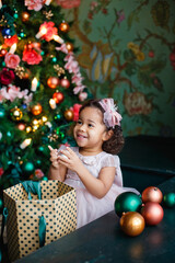 a little African-American girl in a pink dress is playing with Christmas balls to decorate the Christmas tree.