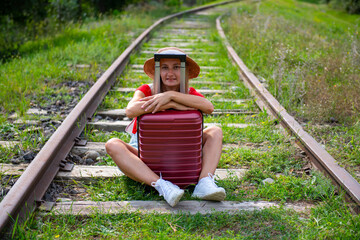 a traveler wearing a hat sits on rails with a suitcase