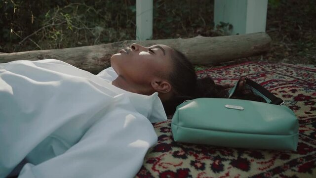 A close-up of a beautiful black woman laying on the red carpet at the forest pose for the camera wearing white trendy jacket and bag.