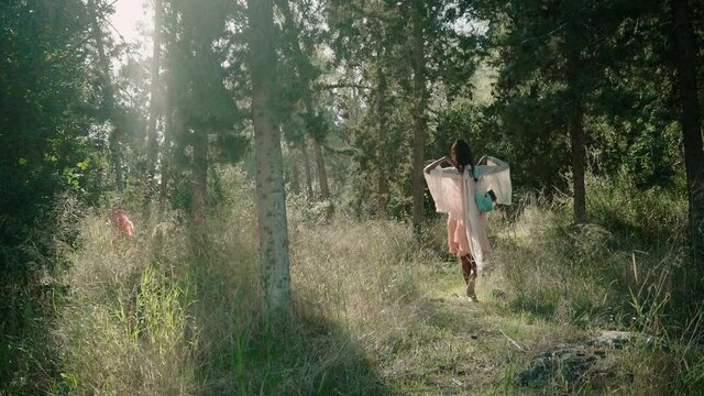 A beautiful black woman walks away from camera with a hand bag as she raises her hands in a beautiful forest