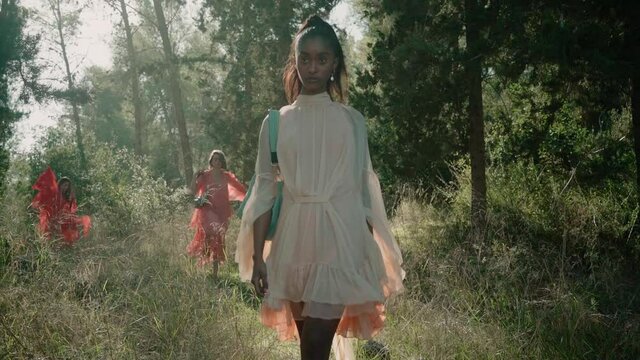 A beautiful black woman walks towards the camera with a hand bag and pass it in a beautiful forest. behind her more models walks