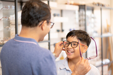 Father hold one of glasses and also touch the other glasses that wearing by his son during selection proper style and type of product in optical shop.