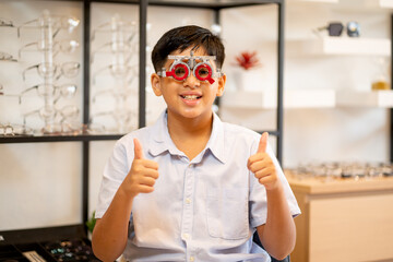 Indian boy wear special test glasses and smile look to camera also show thumbs up with background of many eye glasses on shelves.