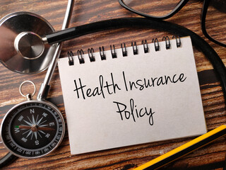 Notebook written with text Health Insurance Policy with Stethoscope,pencil,glasses and compass on a wooden background.