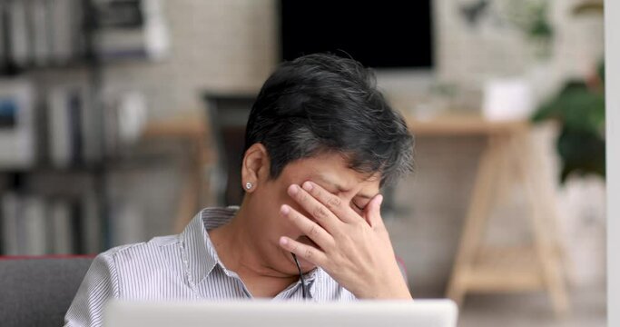 Exhausted mature Asian woman with headache rubbing eyes and touching head while sitting on sofa and working on remote project on laptop at home