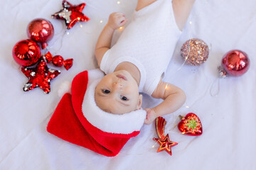 baby in a white bodysuit and a Santa hat is lying on his back on a white sheet surrounded by red...