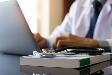 Male doctor working on laptop computer with stethoscope and book on the desk at medical office in clinic or hospital.