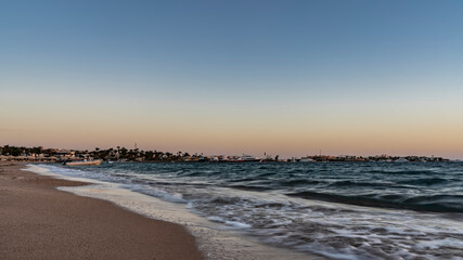 Early morning on the Red Sea beach. Turquoise waves foam on the sandy beach. In the distance you can see sun umbrellas, palm trees, boats. The sky is highlighted in orange. Egypt