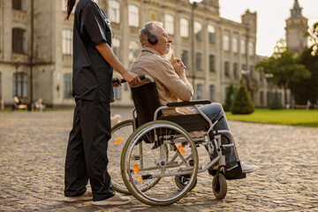 Obraz premium Relaxed senior handicapped man in wheelchair wearing headphones and listening to music on a walk with nurse outdoors