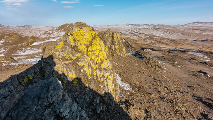 Yellow lichens grow on the cracked slopes of the cliff. The hills in the distance are covered with snow. Blue sky. A sunny winter day. Baikal. Olkhon Island
