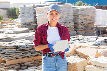 Young positive manager girl working in a building materials store demonstrates a stone tile in an open air warehouse, holding ..it in her hand