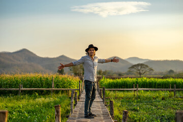 Happy young handsome man and a hat while traveling with glad positive expression in Asia. Attractive male in harmony with the field nature raises his hands to sunset or sunrise and mountain landscape