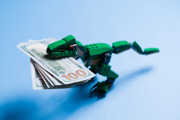 A pack of 100 dollar bills in mouth of tyrannosaur green kid toy isolated on blue background. Copy space. Concept of a unique advertising and design.