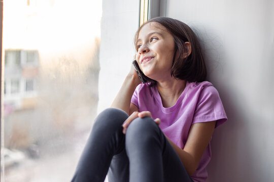 A pre-adolescent girl sitting on a windowsill communicates on the phone with her grandmother or friends while in quarantine.