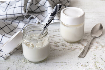 Two jars of homemade yogurt are on the table. One of them is open. A spoon and a towel lie nearby.