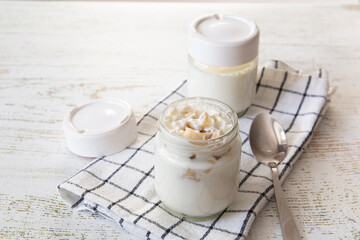 Two jars of homemade yogurt are on the table. One of them is open. A spoon and a towel lie nearby.