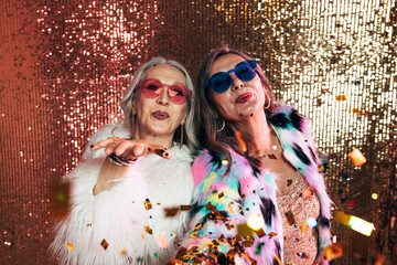 Two senior women in eyeglasses wearing fur coats blowing confetti off their hands against glitter...